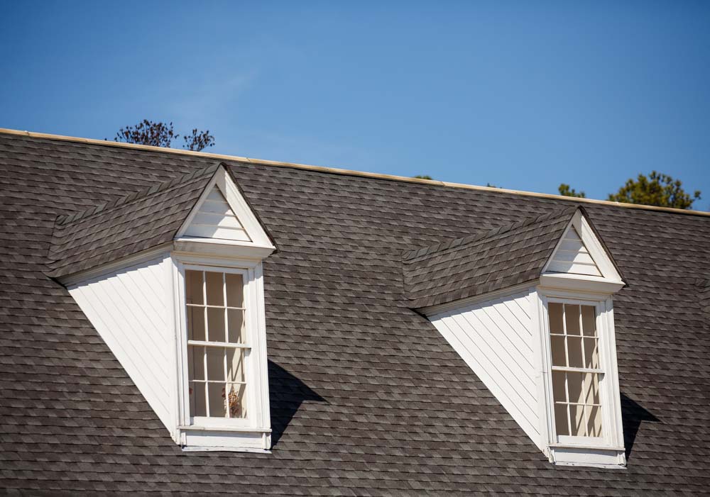 A close-up of a roof with gray shingles and two white dormers.