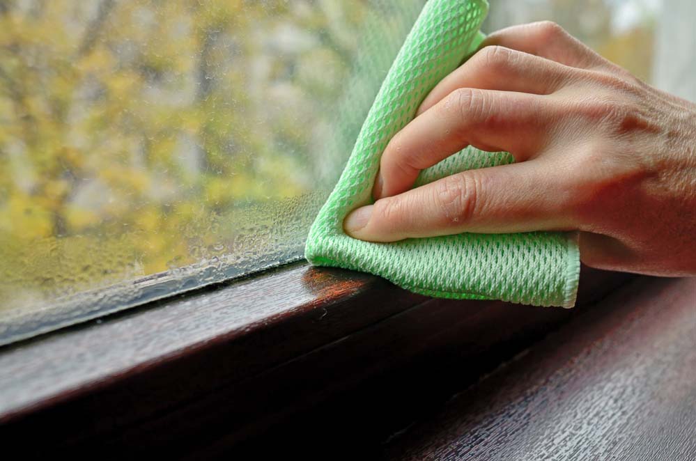A hand uses a green cloth to wipe condensation off the inside pane and sill of a window.
